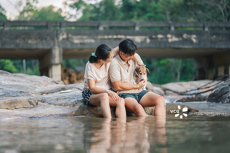 一对老年夫妇在河边与他们的狗互动，展现了人类与宠物之间的深厚情感，背景是宁静的自然环境。图片素材