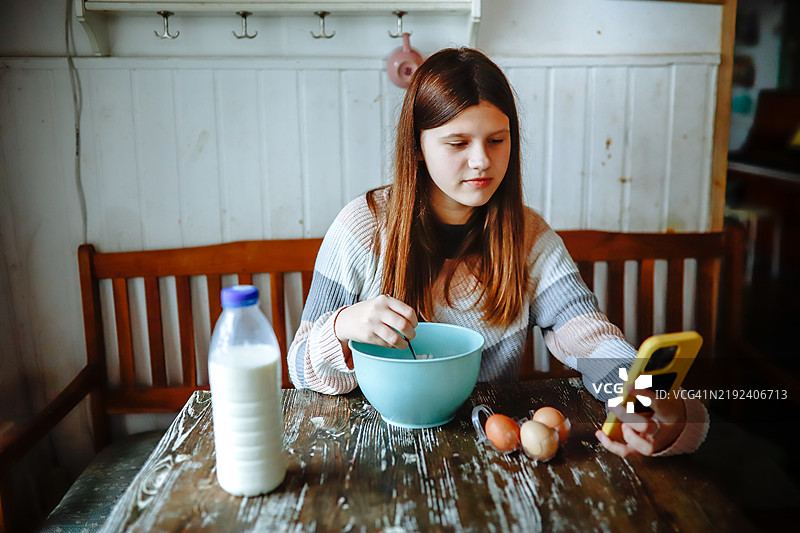 独立烹饪：少女在家中一边拿着智能手机，一边在桌子上搅拌面团和牛奶。图片素材