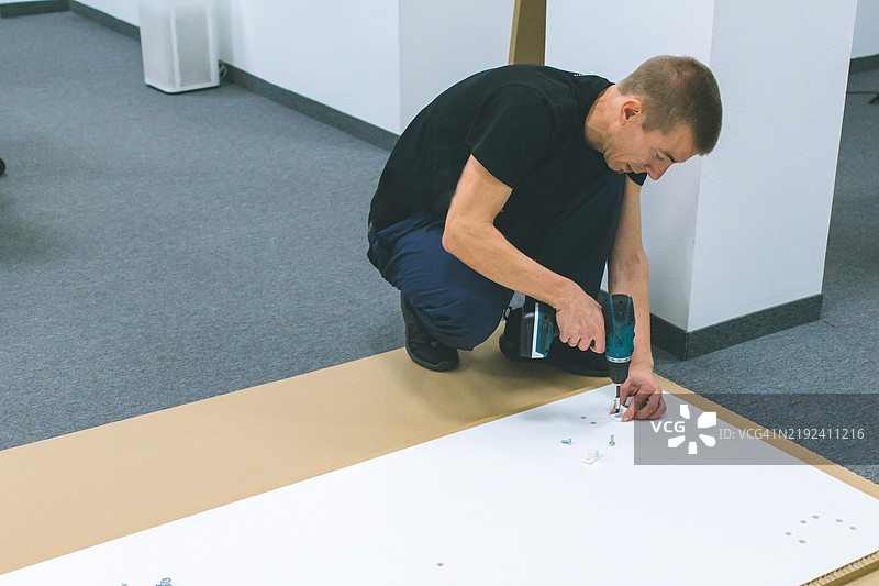 中年工匠在现代办公室工作区的地板上组装家具，使用电动无绳电动工具。图片素材