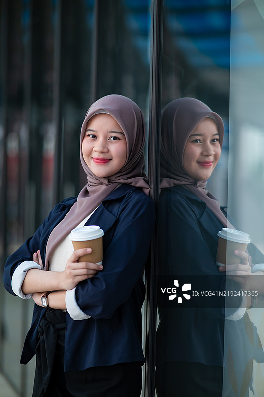 年轻的穆斯林女商人穿着休闲商务装，戴着头巾，在现代办公大楼外享受一杯热咖啡。图片素材
