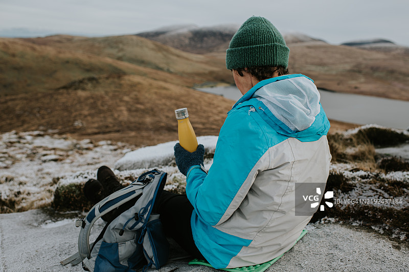 一位活跃的老年人在攀山时休息，停下来坐在冰冷的岩石上享受一杯清爽的饮料。图片素材