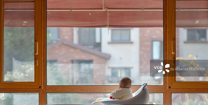 一个亚洲婴儿坐在婴儿椅上，望着窗外，手里拿着玩具，婴儿的母亲在旁边化妆，婴儿六个月大。图片素材