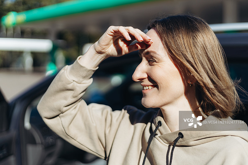 微笑的女性在阳光明媚的日子里站在加油站旁的SUV旁遮挡阳光。图片素材