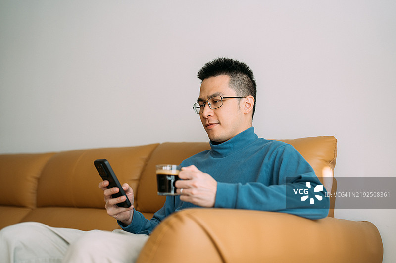 亚洲男子坐在沙发上放松，手里拿着一杯咖啡，正在使用智能手机。图片素材