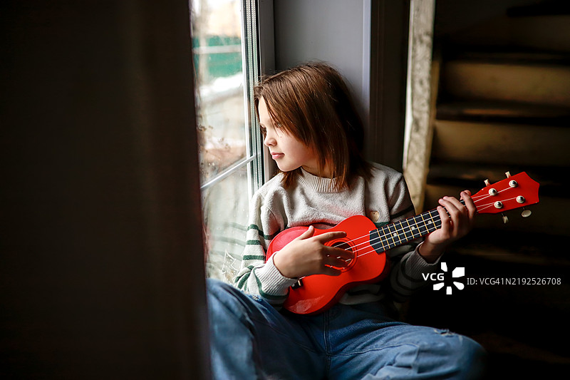 孩子独自在窗台上弹奏尤克里里，表达出在昏暗家居环境中的忧郁。图片素材