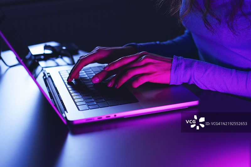 在紫色灯光下的生动黑暗环境中，女性正在笔记本电脑上打字图片素材