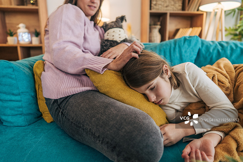 快乐的妈妈坐在青绿色沙发上，使用手机，而她的女儿躺在沙发上，看起来不太舒服。图片素材
