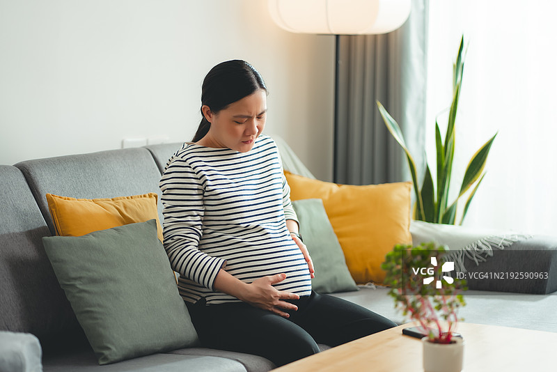 亚洲孕妇坐在家中的沙发上感到腹痛。图片素材
