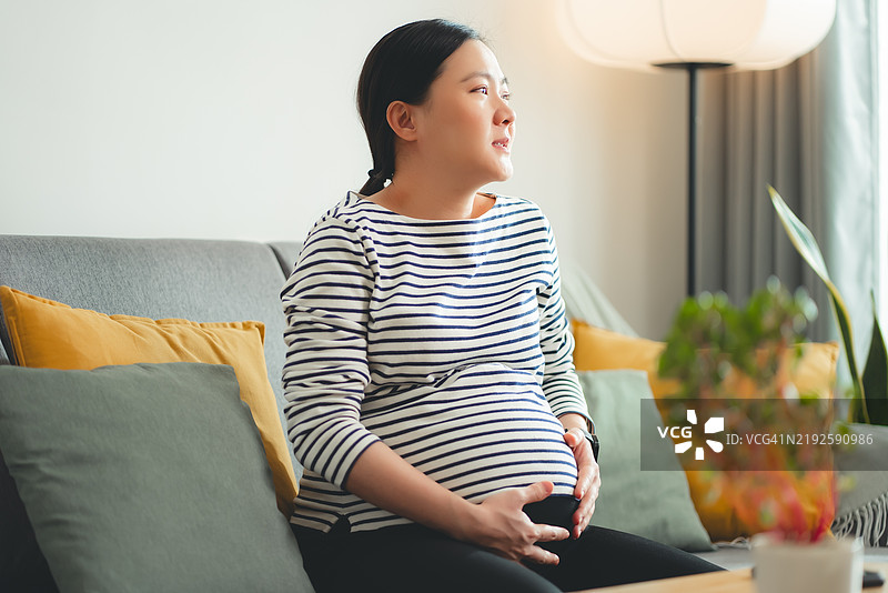 亚洲孕妇坐在家中客厅的沙发上感到腹痛。图片素材