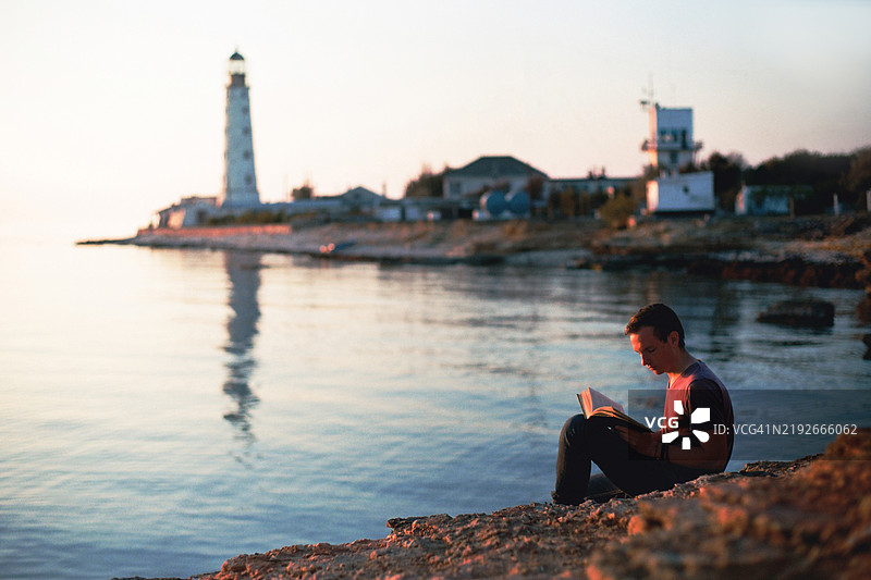 年轻人在灯塔背景下的海岸边阅读书籍图片素材