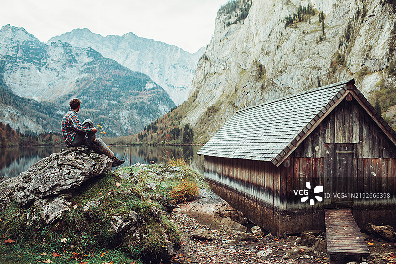 男子在秋季时分，站在巴伐利亚阿尔卑斯山的科尼希湖旁，凝视着木船屋。图片素材