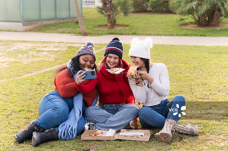 快乐的女性朋友们在公园里吃披萨时自拍图片素材