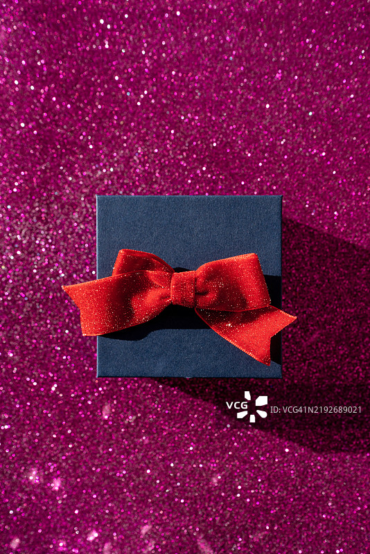 红色蝴蝶结的礼品盒静静地放在鲜艳的粉色闪光背景上，散发出节日的魅力。图片素材