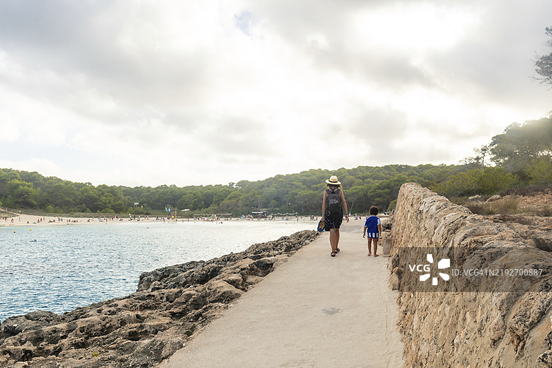 母亲和儿子在海边的小路上散步，享受西班牙马洛卡的萨马拉多海滩的夏日假期。图片素材