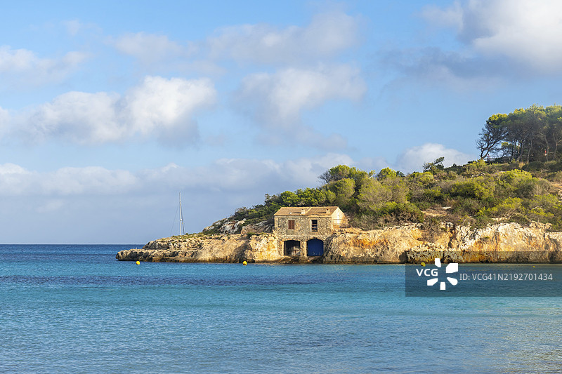 马洛卡的萨马拉多海滩宁静的场景，岩石突出部上有一座石制船屋，碧蓝的海水和远处的帆船相映成趣。图片素材
