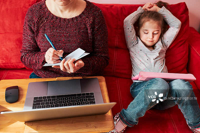 母亲在家通过笔记本电脑进行视频聊天，远程工作，同时她的女儿在旁边用平板电脑观看视频。图片素材