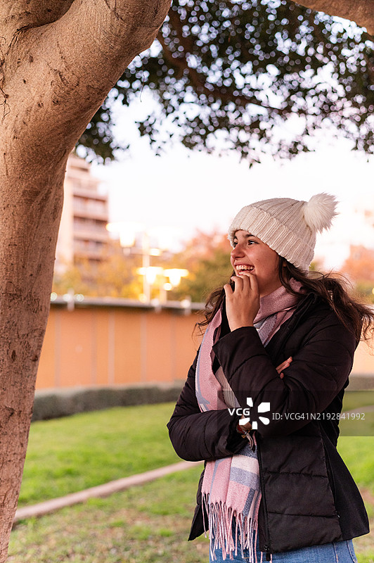 年轻女性在冬季的公园里微笑着倚靠在树上图片素材