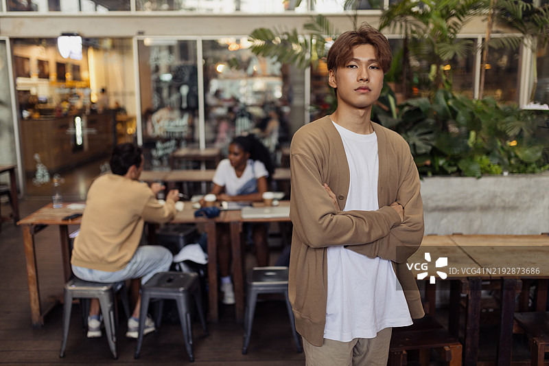自信的年轻男子站在咖啡馆里，背景中与朋友交谈。图片素材