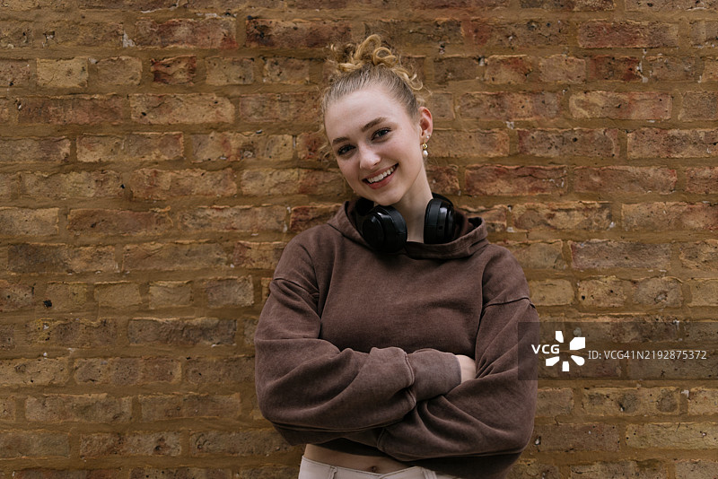 年轻女性在户外微笑，佩戴耳机，背景是砖墙。图片素材