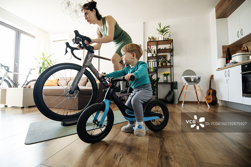 母亲在家里的静止自行车上，旁边有一个骑儿童自行车的幼儿。图片素材