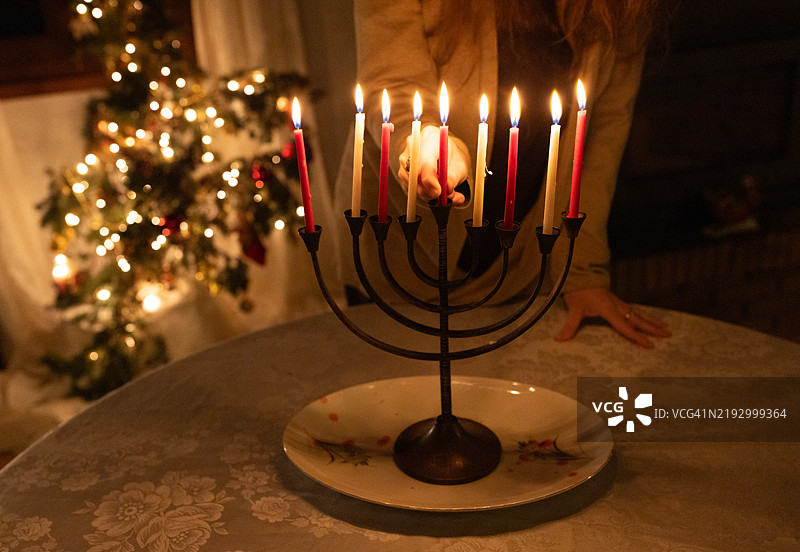 一位女性的手特写，将辅助蜡烛（shamash）放入烛台中，点燃了汉努基亚（hanukkiah）上的蜡烛，庆祝光明节的第八个夜晚。图片素材