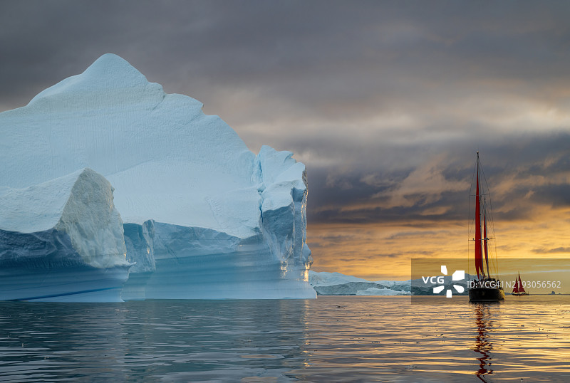 在格林兰伊卢利萨特的迪斯科湾冰川中，一艘红色帆船在漂浮的冰山之间航行，背景是极昼夏季的橙色日落。图片素材