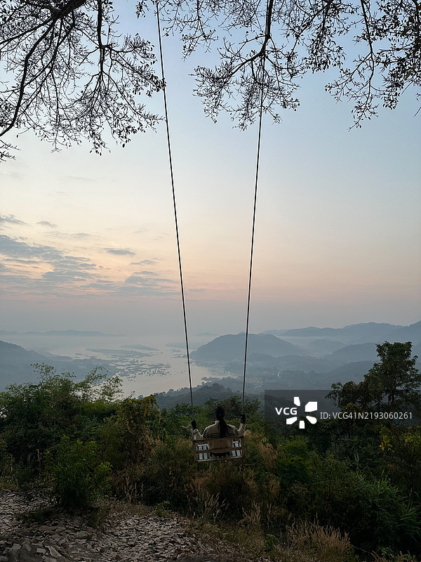 一幅宁静的画面，描绘一位女性在秋千上享受日出时分的美丽山景，四周环绕着郁郁葱葱的绿树。图片素材