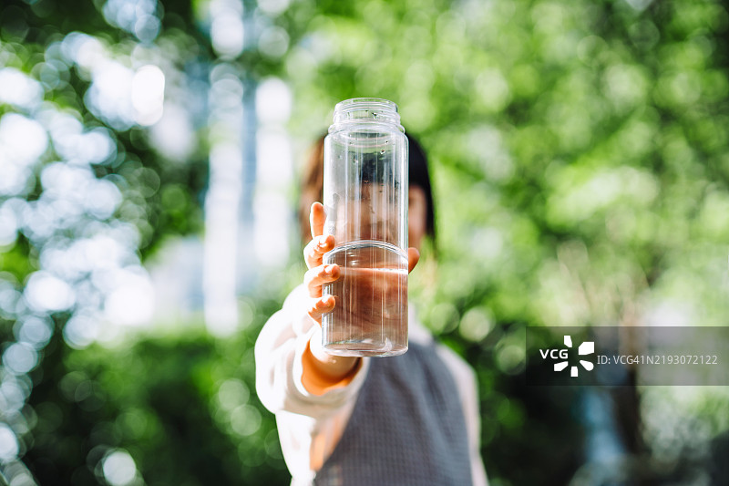 亚洲女孩在阳光下的公园里，手持可重复使用的塑料瓶，遮住脸部喝水，周围是绿色植物。保持水分。健康生活方式。环保、零浪费和可持续生活理念。图片素材