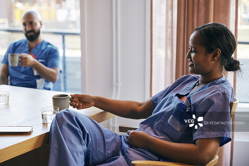 微笑的女护士在医院的咖啡厅里靠在椅子上，手里拿着茶杯，享受咖啡休息时间。图片素材