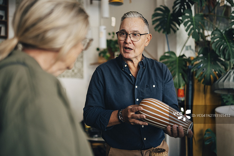 年长男性店主手持花瓶，与正在古董店购物的女性顾客交谈图片素材