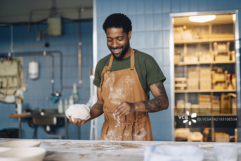 微笑的男性面包师在面包店工作，撒面粉在桌子上并握着面团。图片素材