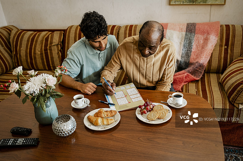 老年男子在家中沙发上解数独，旁边坐着一位男护士。图片素材
