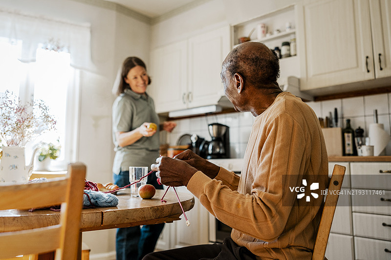 侧面视图的老年男性在家中的厨房里一边编织一边与女性护士交谈图片素材