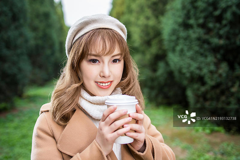 一位穿着棕色外套和白色围巾的年轻女性正微笑着手握一杯咖啡。图片素材
