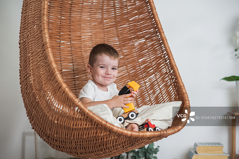 微笑的男孩在悬挂的藤椅上玩玩具车图片素材