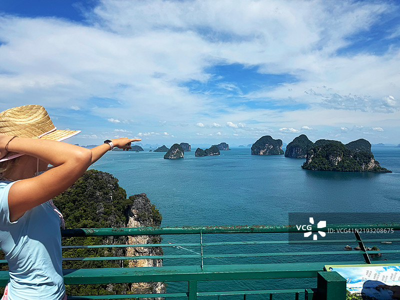 一位美丽年轻女性穿着蓝色T恤和草帽，站在泰国岛的观景平台上，指向海中的岛屿。周围是美丽的岩石风景。热带假期。图片素材