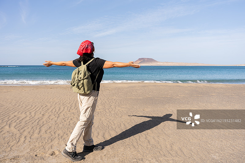 女性徒步旅行，张开双臂，拥抱火山岛的全景，背景是阳光明媚的海滩。图片素材