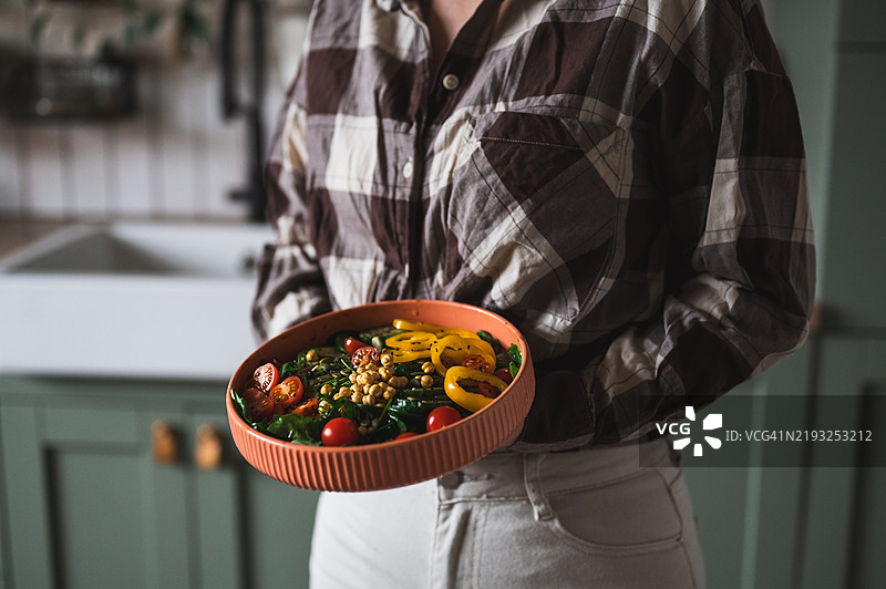 健康的晚餐或午餐。穿着T恤和牛仔裤的女性站着，手中拿着装有鹰嘴豆泥、蔬菜、沙拉、豆类、库斯库斯和鳄梨的素食超级碗或佛碗，以及一杯冰沙，方形裁剪。图片素材