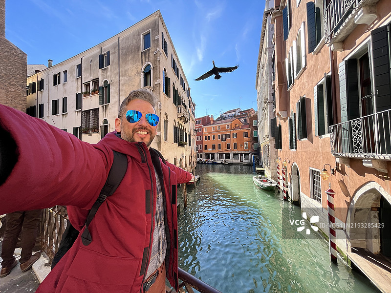 游客在威尼斯拍摄自拍，头顶有一只鸬鹚飞过运河。图片素材
