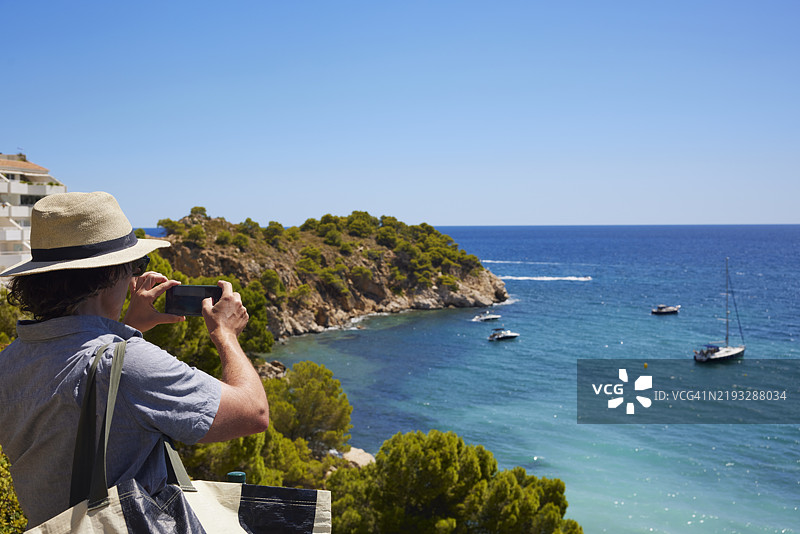 男子在西班牙白色海岸的马斯卡拉特海滩拍摄海湾、碧绿海洋中的船只和岬角图片素材