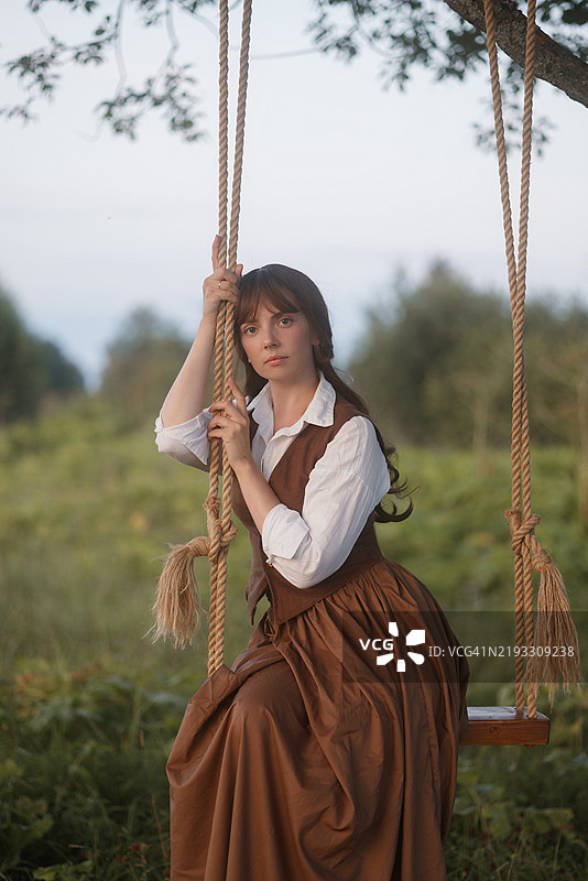 穿着棕色复古裙子的年轻女性在田野中的粗绳秋千上。图片素材