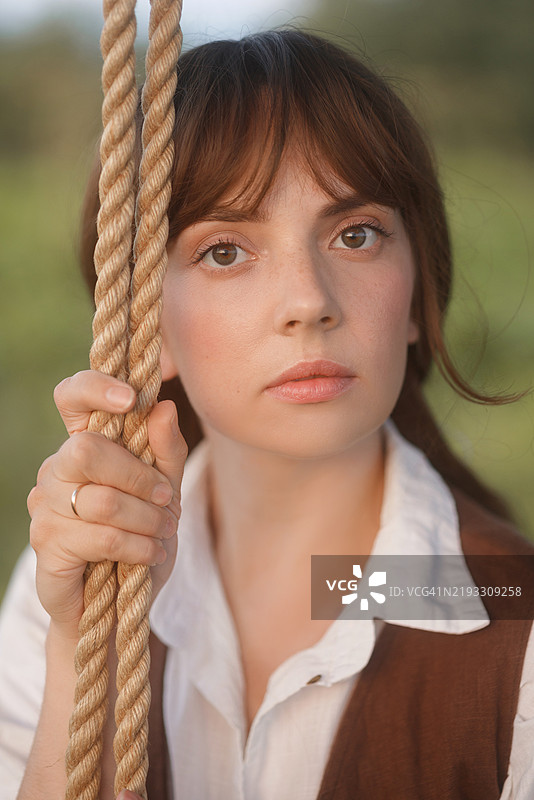 穿着棕色复古裙子的女人在田野中的粗绳秋千上特写肖像。图片素材