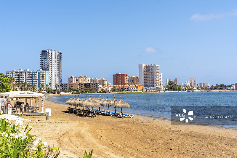 宁静的海滩场景，配有草编伞和躺椅，俯瞰西班牙穆尔西亚的马尔门诺尔地区拉曼加的城市景观，卡瓦纳海滩图片素材