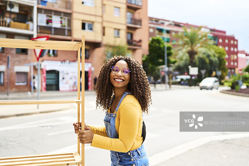 美丽的棕发女性佩戴眼镜，站在街上微笑着看向镜头，侧面视图，手里拿着一个木架子。图片素材