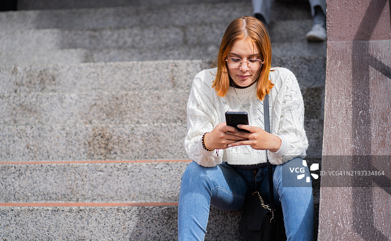 一位美丽的女性坐在街道上的台阶上，手里拿着智能手机 - 股票照片图片素材