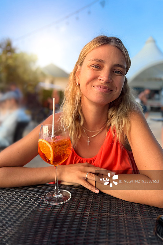 年轻美丽的女人穿着红色连衣裙，在海滩酒吧的日落时分微笑着喝着阿佩罗鸡尾酒。图片素材