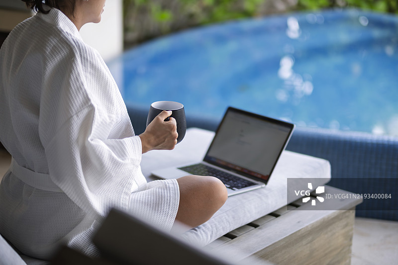 度假中的女性在泳池前一边喝咖啡一边使用电脑工作图片素材
