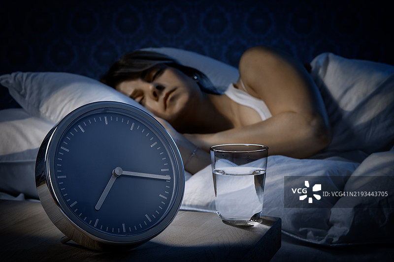夜晚，女人在床上睡觉，旁边有响铃的闹钟和一杯水。图片素材