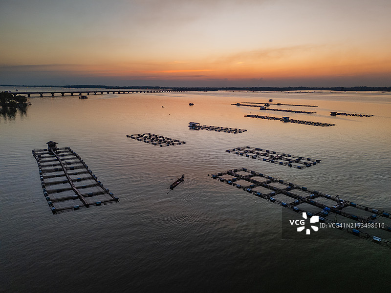 宁静的空中视角，夕阳下平静湖面上的漂浮鱼塘，远处有一座横跨地平线的桥。图片素材