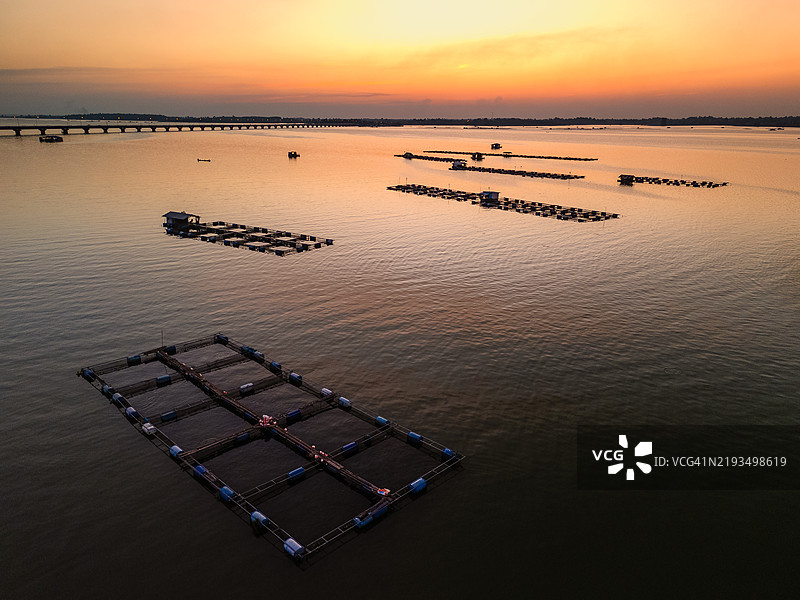 宁静的空中视角，夕阳下平静湖面上的漂浮鱼塘，远处有一座横跨地平线的桥。图片素材